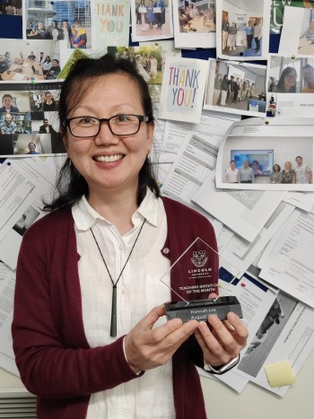 Hannah Lee with her award and standing in front of a notice board full of photos and cards from past students